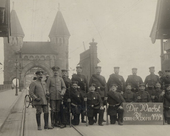 Das Schwarz-Weiß-Foto zeigt Männer in Uniform an einer Brücke. Sie halten ein Schild, auf dem steht: Die Wacht am Rhein. Im Hintergrund ist ein Torbogen der Brücke.