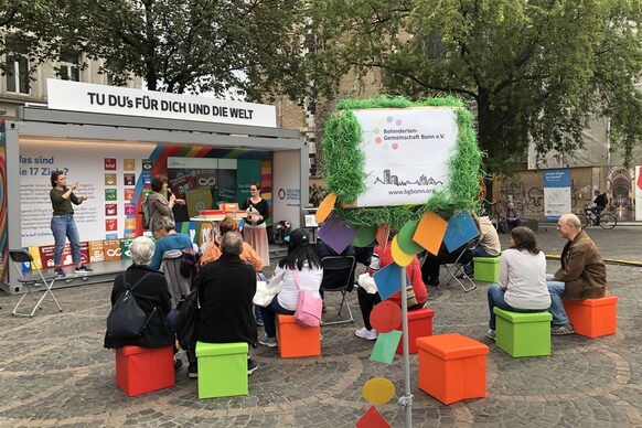Mehrere Personen sitze auf orangen und grünen Kisten auf einem Platz vor einem Infostand