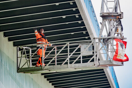 Ein Mann in orangefarbener Schutzkleidung kontrolliert im Hängekorb die Unterkonstruktion der Kennedybrücke