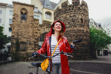 Eine Frau in rotem Regenmantel steht mit ihrem Fahrrad vor dem Bonner Sterntor im Regen und lacht.