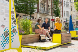 Drei Menschen sitzen auf einem mit Grün bepflanztem Stadtmöbel und unterhalten sich; links daneben ist eine Informationstafel.
