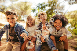 Vier lachende Kinder sitzen auf einem Baumstamm und pusten Seifenblasen