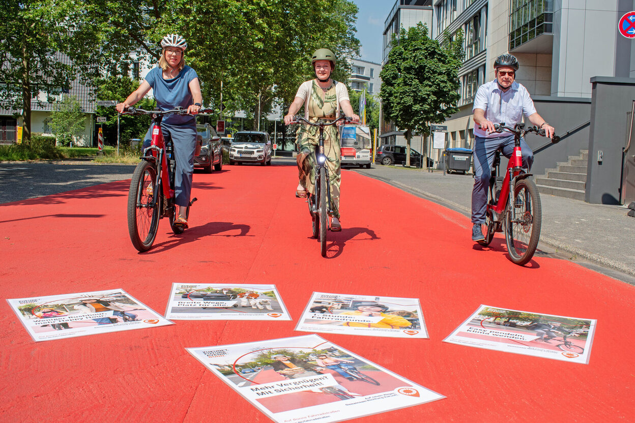 Auf Fahrrädern in der neuen, rot markierten Fahrradstraße in der Fritz-Erler-Straße: (v.r.) Stadtbaurat Helmut Wiesner, Oberbürgermeisterin Katja Dörner und Stadtsprecherin Barbara Löcherbach. Die auf dem Boden liegenden Plakatmotive werden auf digitalen Informationstafeln im Stadtgebiet gezeigt.