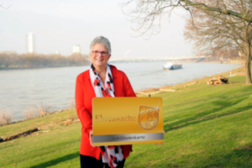 Das Bild zeigt Monika Fritzsche, die am Beueler Rheinufer steht und eine symbolische Jubiläums-Ehrenamtskarte in den Händen hält.