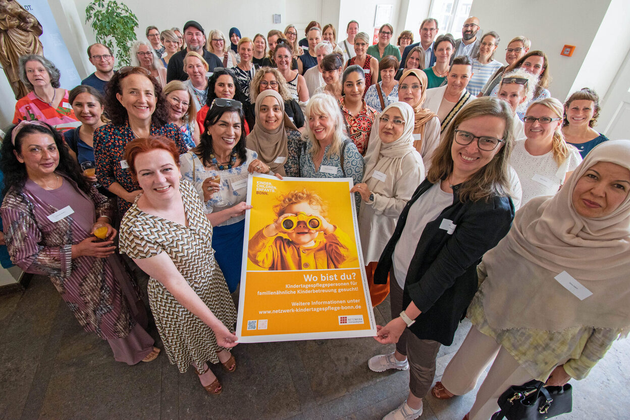Das Bild zeigt Oberbürgermeisterin Katja Dörner und die geladenen Kindertagespflegepersonen beim Empfang im Alten Rathaus. Gemeinsam zeigen sie das Plakat der neuen Kampagne zur Gewinnung von neuen Tagespflegepersonen.