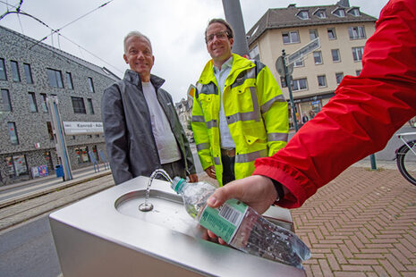 Am neuen Trinkwasserbrunnen am Konrad-Adenauer-Platz wird eine Flasche befüllt, im Hintergrund stehen zwei Personen.