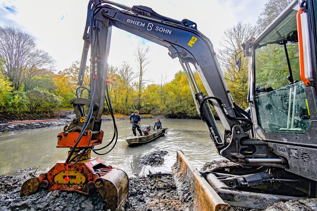 Ein Bagger vor einem schlammigen Teich, im Hintergrund ein Boot mit zwei Menschen.