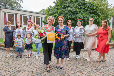 Das Bild zeigt (vorne) Yevheniia Burlaka, die von Oberbürgermeisterin Katja Dörner die 3.000 Ehrenamtskarte NRW erhält. Im Hintergrund sind Mitglieder des Chores Namysto, der Lukaskirchengemeinde und der Bonner Freiwilligenagentur zu sehen.