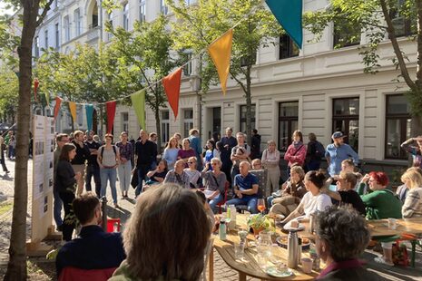 Festlich geschmückte Straße mit Bürger*innen, die im Freien einem Vortrag lauschen.