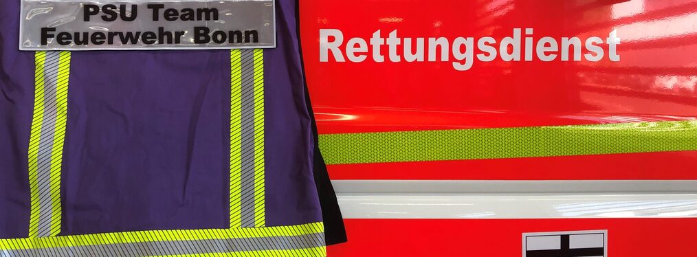 Feuerwehrjacke mit Aufschrift „Teamleiter PSU-Team Feuerwehr Bonn“, im Hintergrund ein Fahrzeug des Feuerwehr- und Rettungsdienstes.